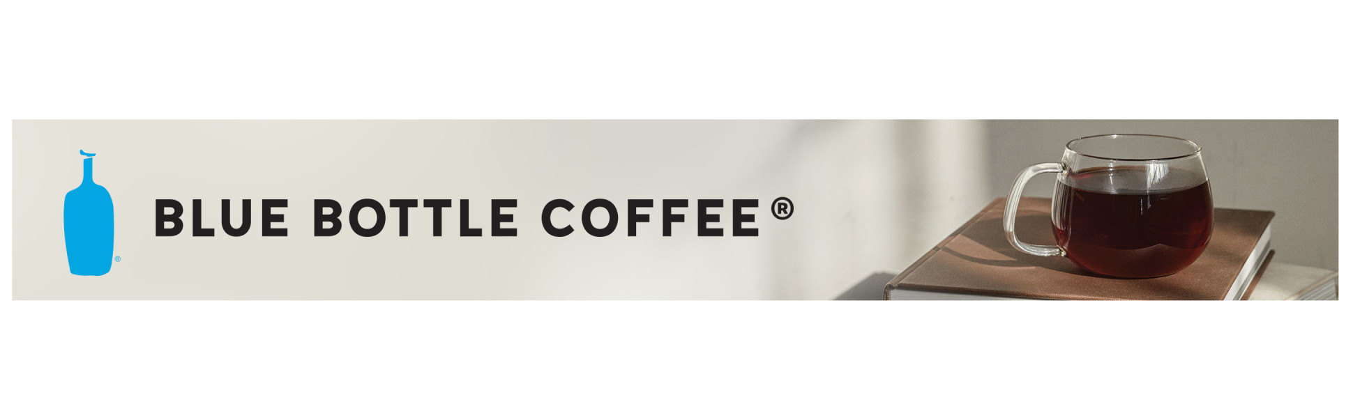 A sunlit, clear glass mug of black coffee on a book. Tagline: Blue Bottle Coffee