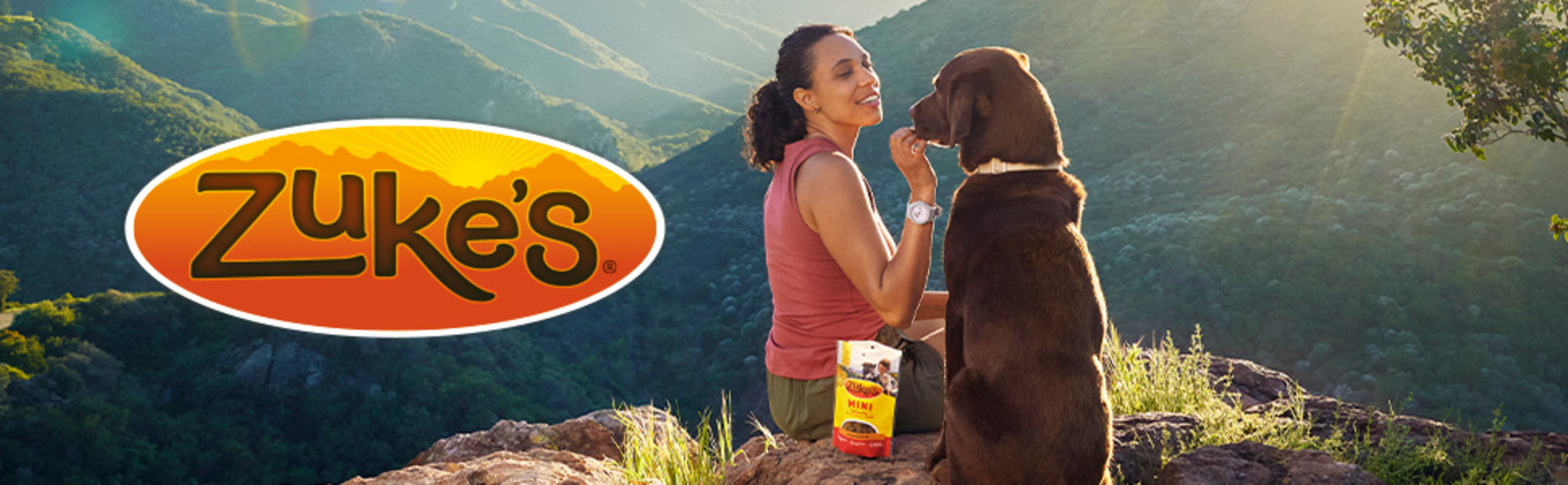 Image of woman giving a dog a treat on a trail overlook. Zuke's logo visible