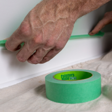 Painters Mate painters tape being applied to a white baseboard in 1 to 2 foot sections. A roll of green tape sits on the floor on top of a protective sheet for painting.