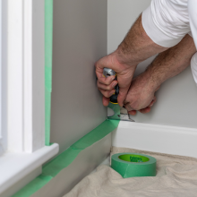 Painters Mate painting tape on a white baseboard and trim because burnished. A roll on green tape is laying on the floor on top of a sheet to protect the floor.