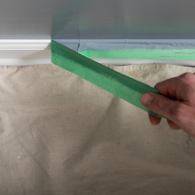 Painters Mate Painters Tape being removed from a white baseboard, revealing the clean paintline between the baseboard and recently painted grey wall.