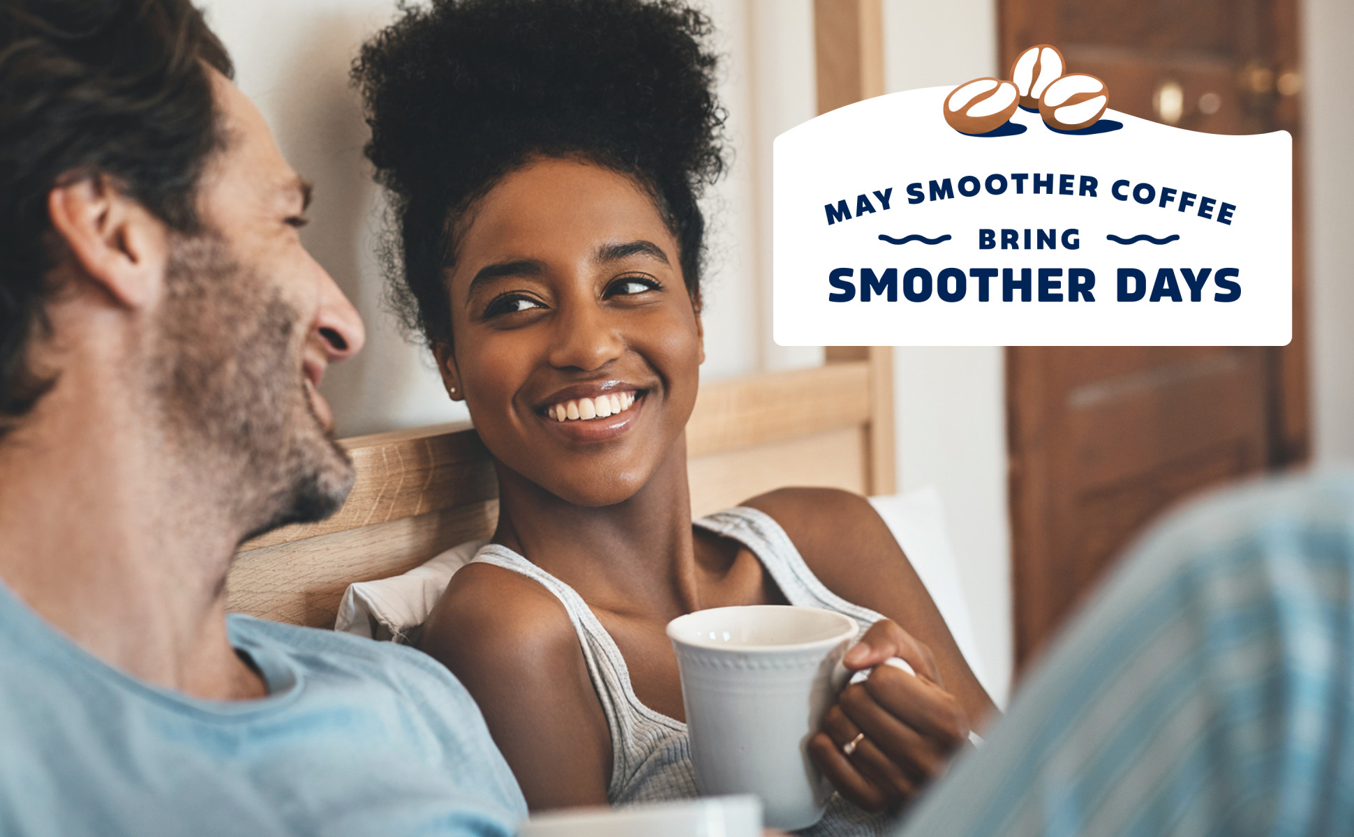 A couple in bed smiling at each other holding coffee mugs. May smoother coffee bring smoother days.