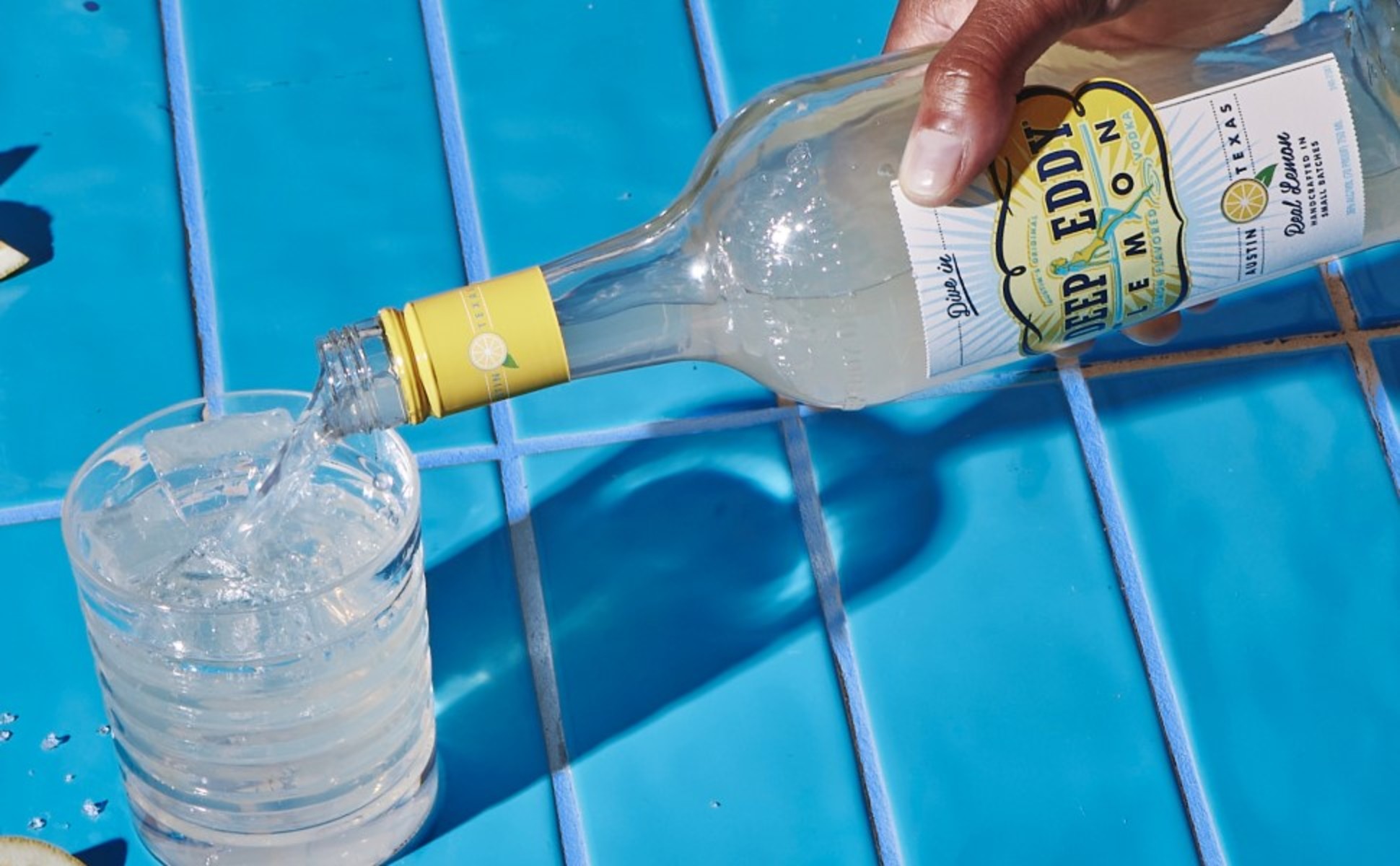 Hand pouring Deep Eddy Lemon Vodka into a glass casting a long shadow on blue tile.