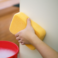 A hand holds a yellow spong washing a wall prior to painting. A red bucket filled with water and soap sits on the floor next to the wall.