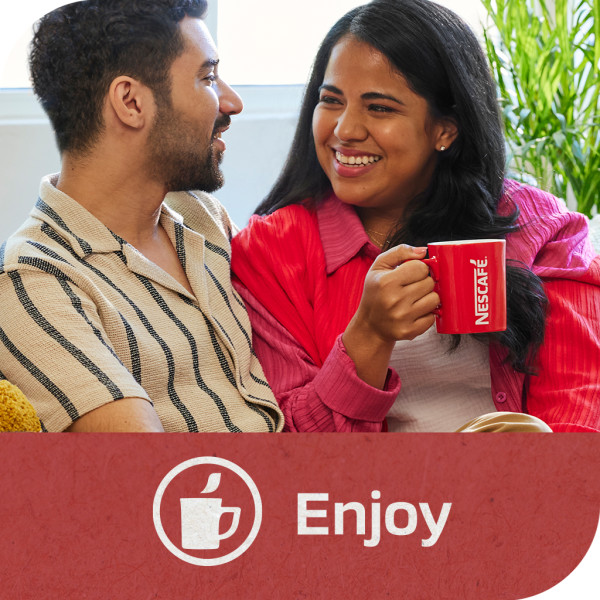 A smiling woman holding a NESCAFÉ mug, sitting with her partner, listening to him. Tagline: Enjoy.