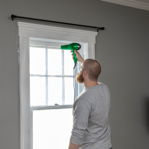 a man using a hair dryer to get the wrinkles out of a shrink film plastic window kit