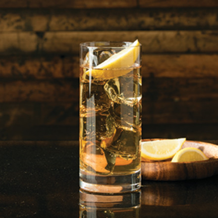 Cocktail in a highball glass with a lemon garnish sitting on a dark counter
